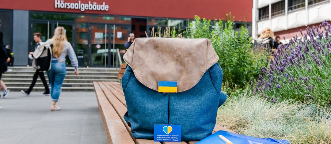 Symbolbild: Auf dem Uni-Innenhof steht ein Rucksack auf einer Bank. Auf dem Rucksack ist eine kleiner Sticker in blau/gelb den ukrainischen Nationalfarben angebracht. Im Hintergrund sieht man verschwommen Menschen in das Hörsaalgebäude gehen.