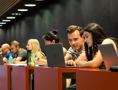 Studierende sitzen zusammen an den Arbeitsplätzen im Hörsaalgebäude und schauen in einen Laptop.