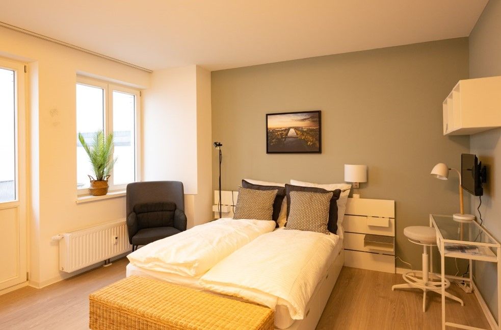 enlarge the image: Cozy furnished bedroom with a double bed, a green-painted accent wall, an armchair by the window, a small desk, a wall-mounted TV with shelves, and a decorative wall picture above the bed. Photo: Thomas Biermann