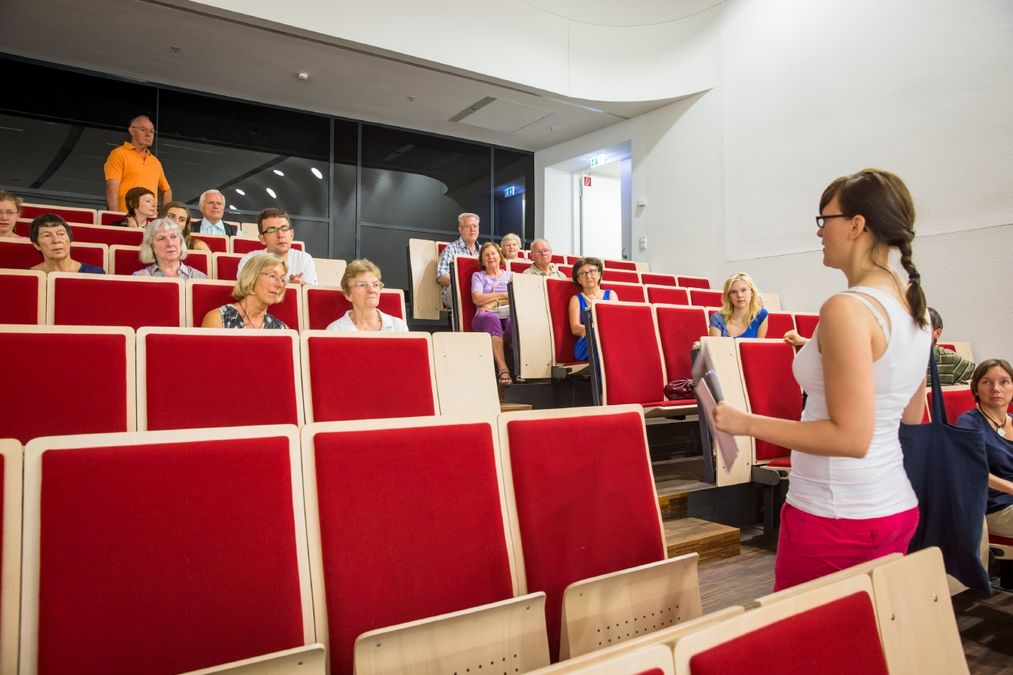 zur Vergrößerungsansicht des Bildes: Foto: eine Gästführerin spricht vor einer Gruppe von Gästen, die in einem Hörsaal Platz genommen haben