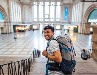 Farbfoto: Eine Person mit Rucksack lächelt in die Kamera. Im Hintergrund befindet sich die Eingangshalle des Leipziger Bahnhofs.