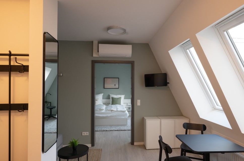 enlarge the image: Cozy apartment with green accent walls, two large skylights, a separate bedroom with a double bed, a small dining table with two chairs, a wall-mounted TV, air conditioning, and a large wall mirror. Photo: Thomas Biermann