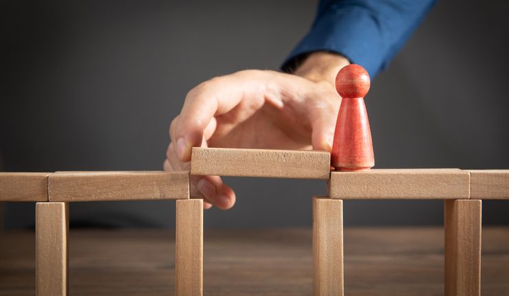 Farbfoto mit Bauklötzen und einer roten Spielfigur. Eine Hand legt einen Bauklotz als Brücke zwischen die anderen, damit die Spielfigur darüber gehen kann.